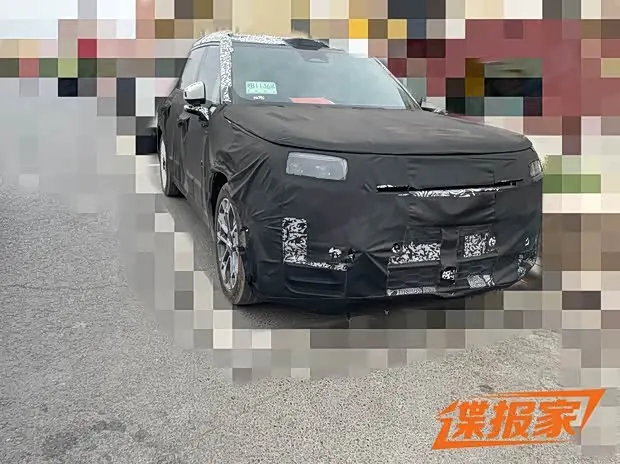 Тестовый Lynk & Co 900 в чёрном камуфляже, вид спереди и слева, на асфальтовой площадке
