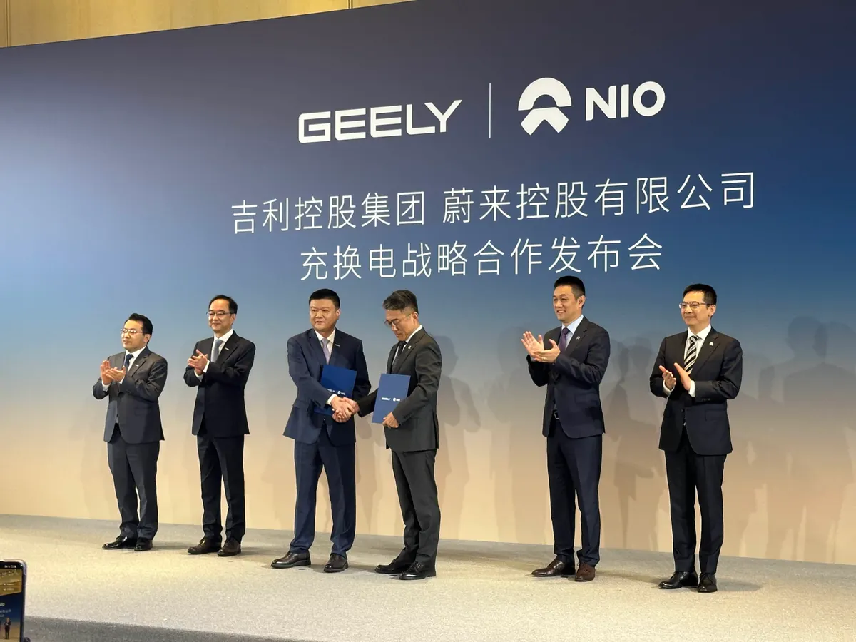 Руководители Geely и NIO пожимают друг другу руки на сцене с папками, рядом аплодируют коллеги