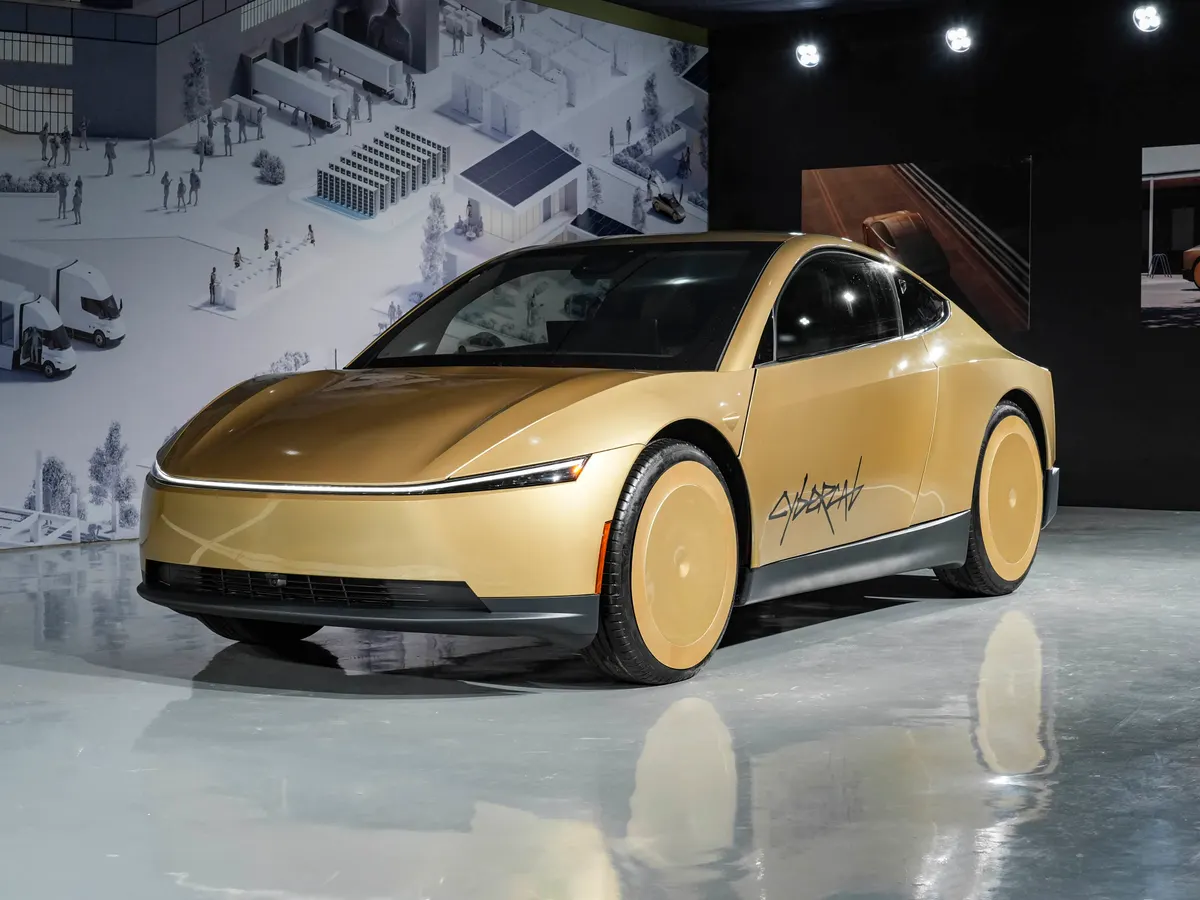 Золотистый электромобиль Tesla на выставке, вид спереди: тонкая полоса фар, закрытые колёсные диски