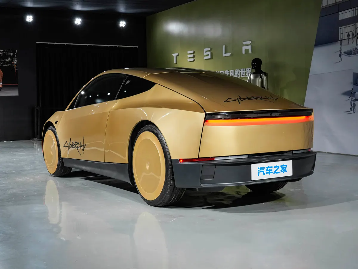 Золотистый Tesla сзади: сплошная красная полоса задних огней, рядом стена с логотипом и робот