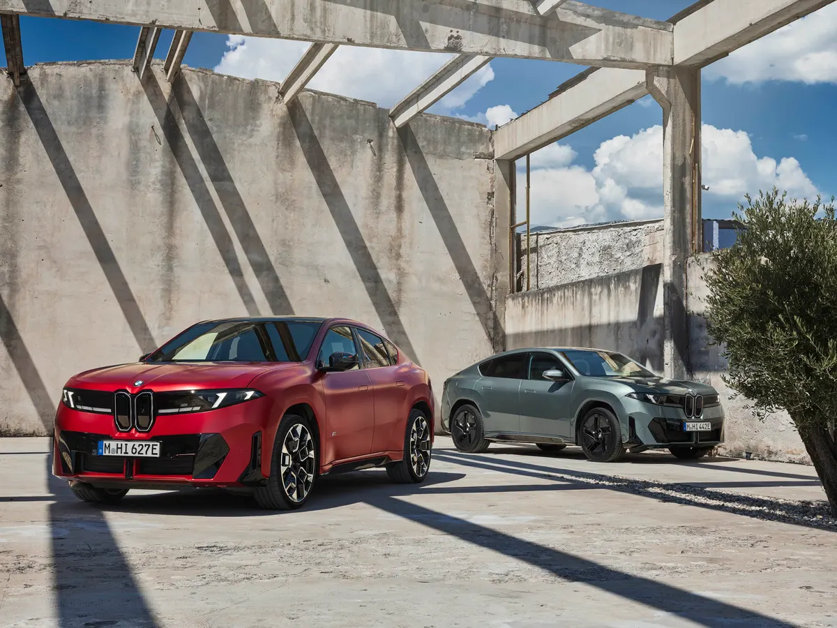 Красный и серо-зелёный BMW iX4 стоят под бетонными балками заброшенного здания на фоне голубого неба