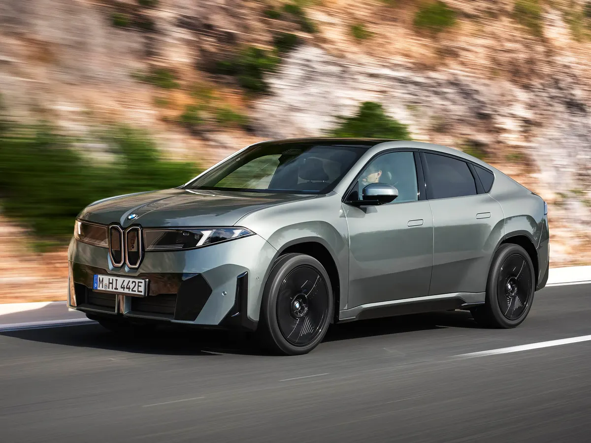 Серо-зелёный BMW iX4 едет по дороге на фоне размытой скалы, вид спереди