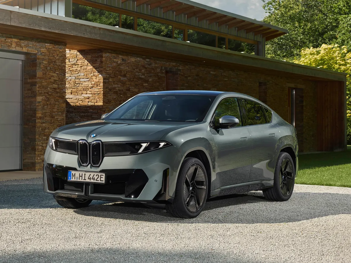 Серо-зелёный BMW iX4, вид спереди в три четверти, у дома с каменной стеной на гравийной площадке