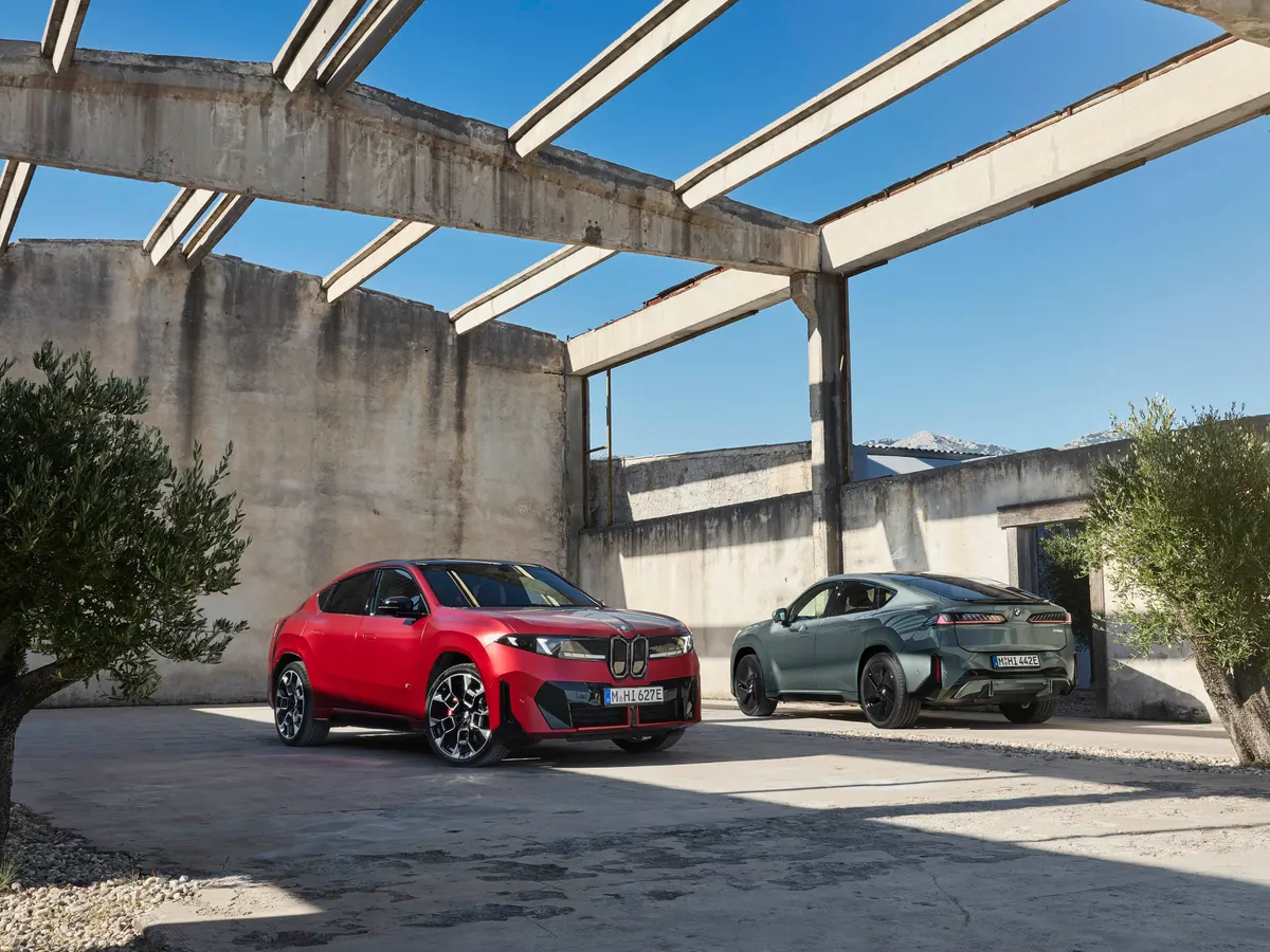 Красный и серо-зелёный BMW iX4 стоят под бетонными балками, рядом оливковые деревья