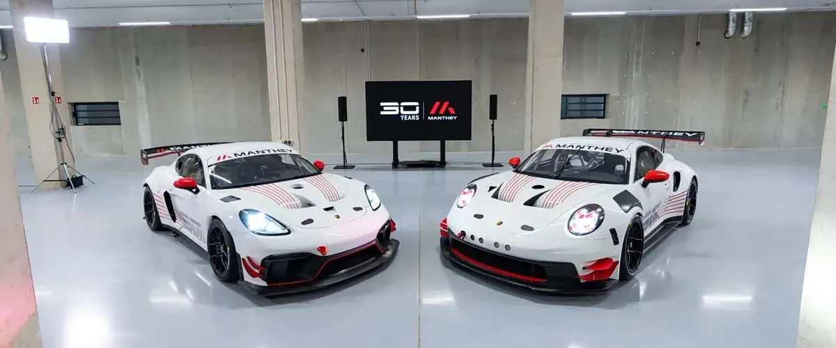 Два белых гоночных Porsche с красными акцентами и надписью Manthey стоят в бетонном зале перед экраном «30 Years»
