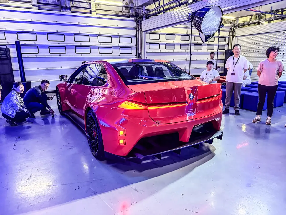 Красный BMW M Concept Neue Klasse сзади сбоку: светящиеся задние огни и люди рядом с машиной