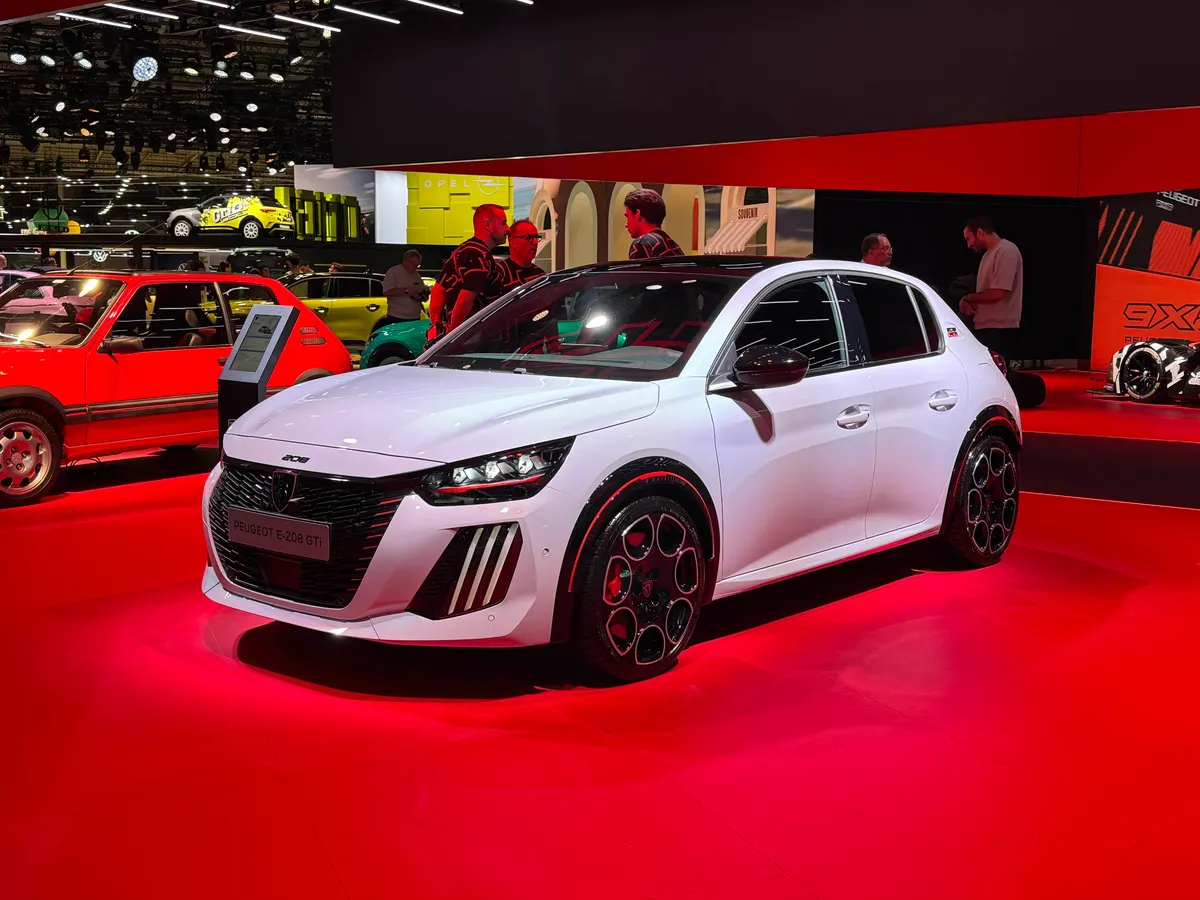 Белый Peugeot E-208 GTi с красной окантовкой арок и чёрными дисками на красной сцене выставки, спереди слева