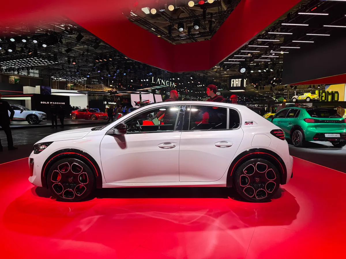 Белый Peugeot E-208 GTi в профиль на красном подиуме, за ним стенды Lancia и Fiat