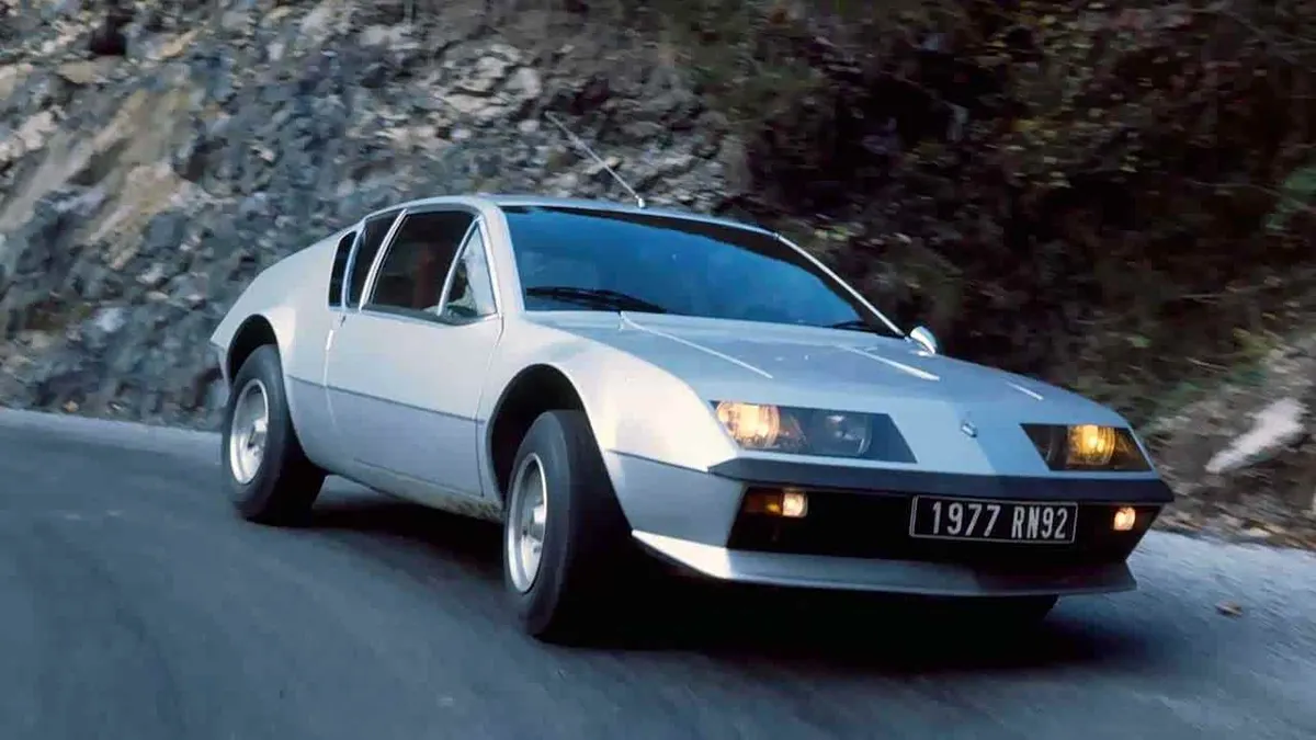 Классическая серебристая Alpine A310 с включёнными жёлтыми фарами на горной дороге, номер 1977 RN92