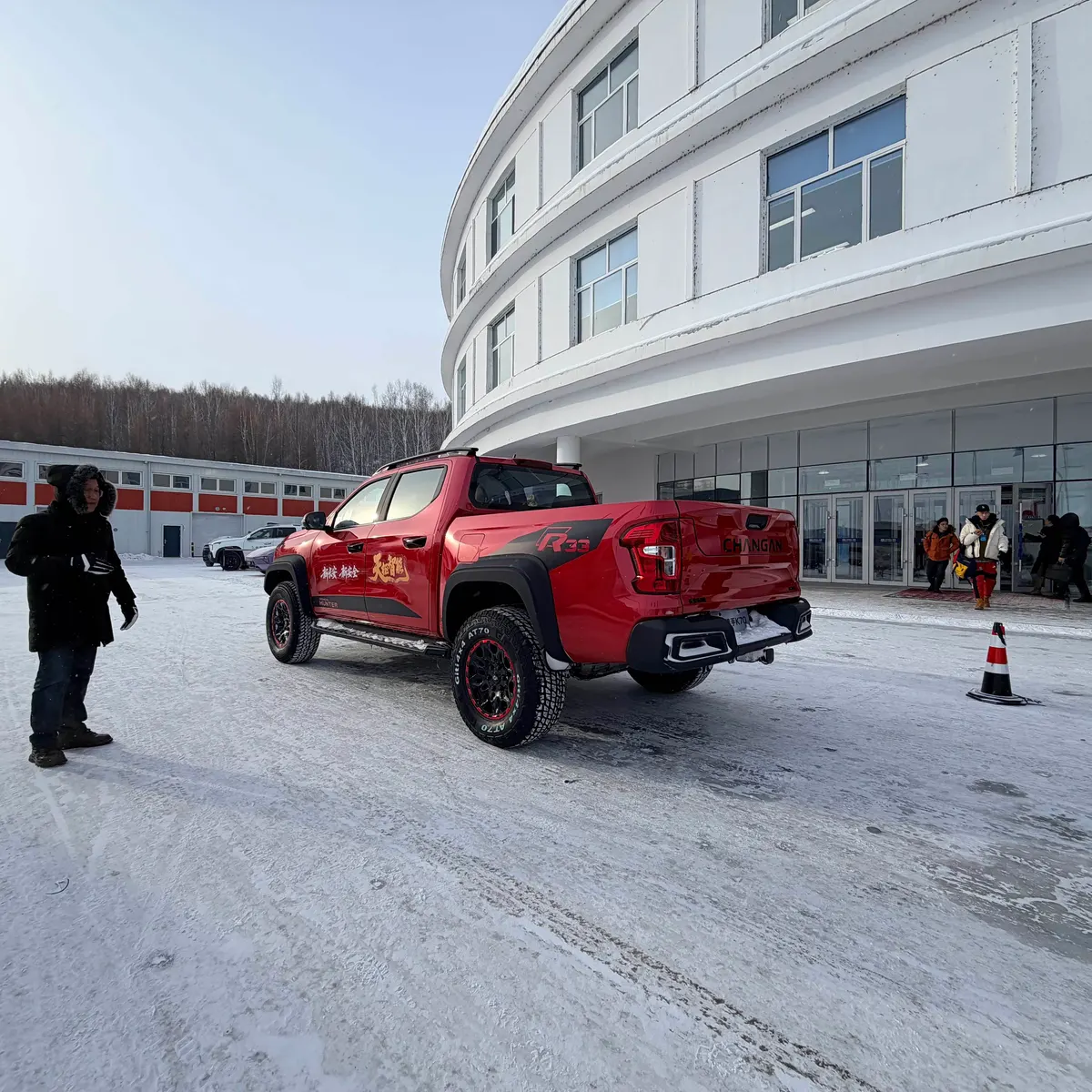 Красный пикап Changan Hunter K70, вид сзади, на снегу у круглого белого здания; слева стоит человек
