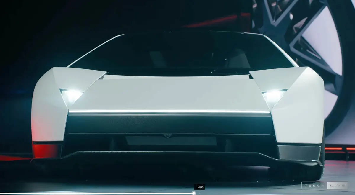 Roadster спереди на сцене: угловатые крылья, треугольные фары, светящаяся полоса, значок Tesla Live