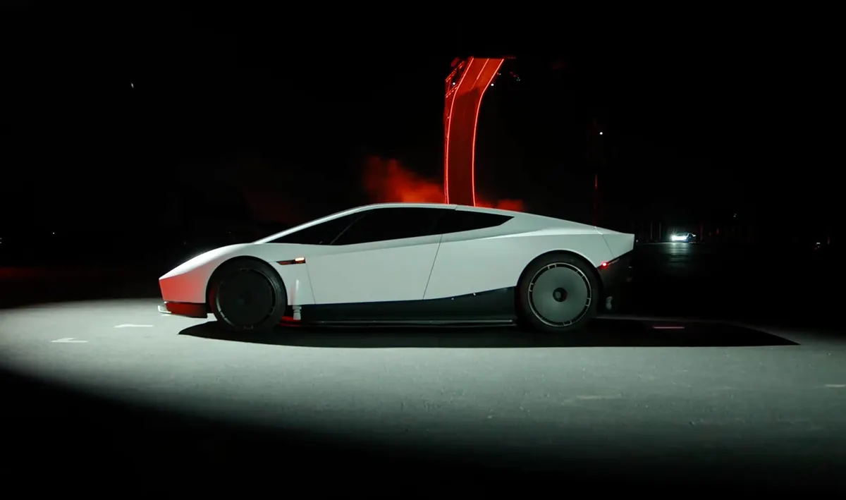 Белый Roadster сбоку на тёмной площадке, позади светится красная рампа