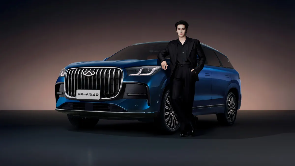 Синий внедорожник Chery Tiggo 9 нового поколения спереди: хромированная решётка, рядом стоит мужчина в чёрном костюме