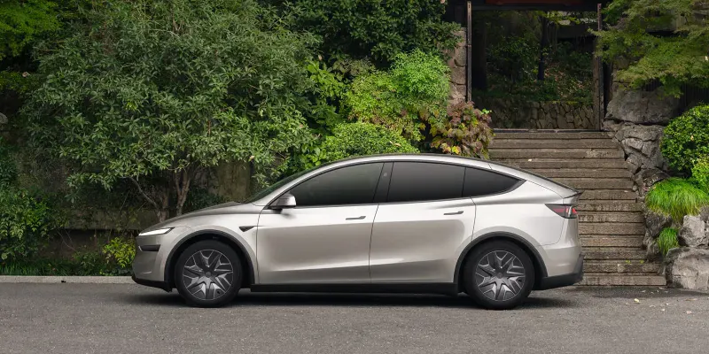 Серебристая Tesla Model Y L сбоку на фоне каменной лестницы и густой зелени