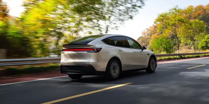Белая Tesla Model Y L в движении, вид сзади, на шоссе среди осенних деревьев