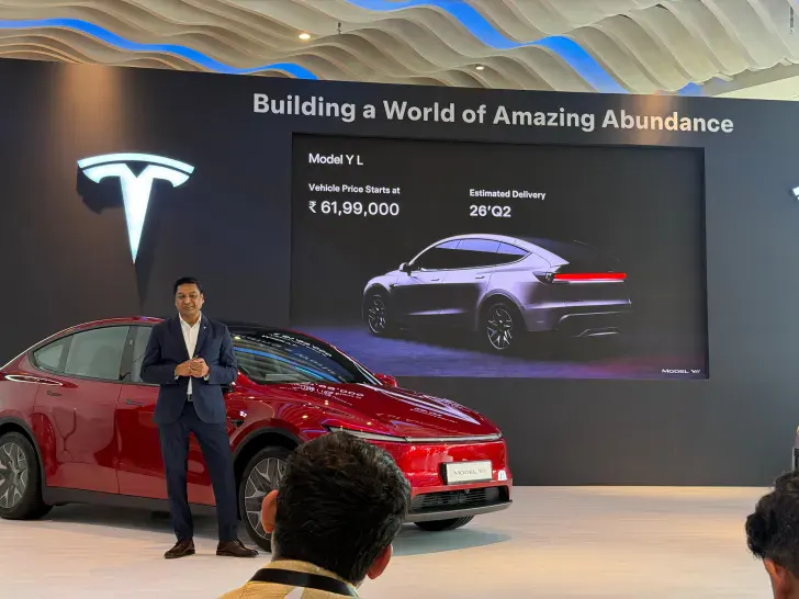 Ведущий в костюме рядом с красной Tesla Model Y L, на экране цена от ₹61,99,000