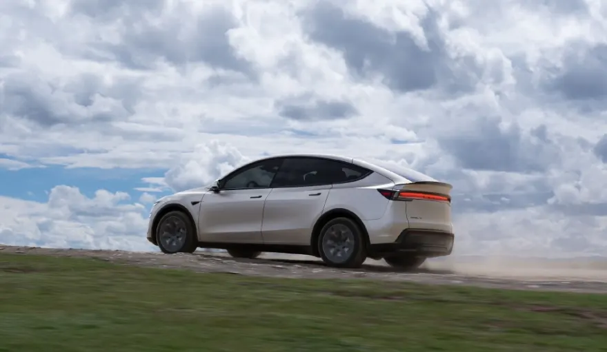 Серебристый кроссовер Tesla едет по грунтовой дороге, поднимая пыль, на фоне облачного неба
