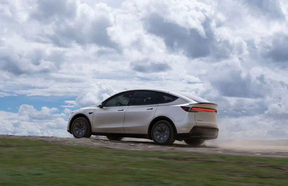 Светлый кроссовер Tesla едет по грунтовой дороге на фоне облачного неба, позади поднимается пыль