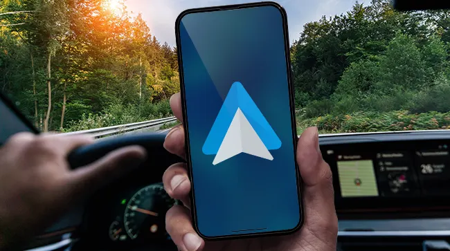 Рука держит смартфон с логотипом Android Auto на синем экране на фоне салона, руля и дороги среди леса
