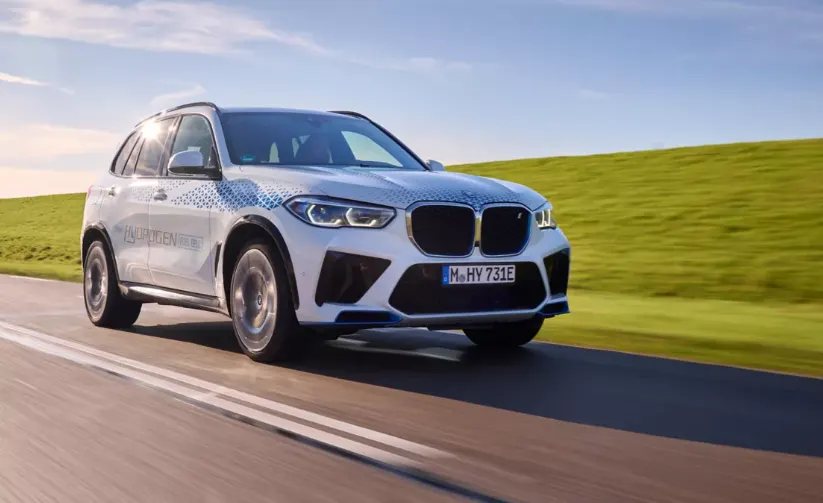 Белый кроссовер BMW iX5 Hydrogen с камуфляжной плёнкой едет по дороге рядом с зелёным полем