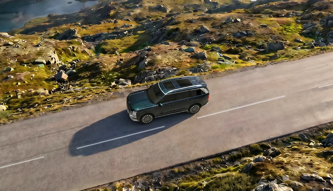 Тёмно-зелёный внедорожник Hyundai, вид сверху, едет по асфальтовой дороге среди каменистых холмов с мхом и озером