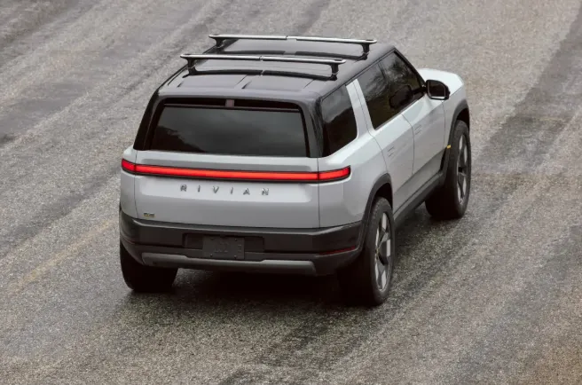 Светло-серый Rivian R2 с чёрной крышей и багажником на крыше, вид сзади сверху, на заднем борту красная световая полоса