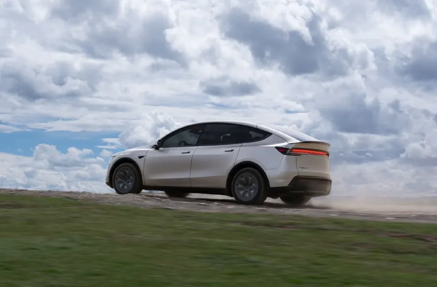 Светлый кроссовер Tesla Model Y едет по грунтовой дороге на фоне облачного неба, за ним поднимается пыль