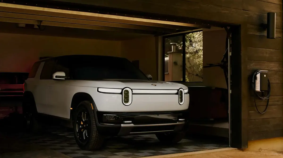 Белый внедорожник Rivian R2 с вертикальными светодиодными фарами стоит в гараже с деревянной отделкой рядом с настенной зарядной станцией