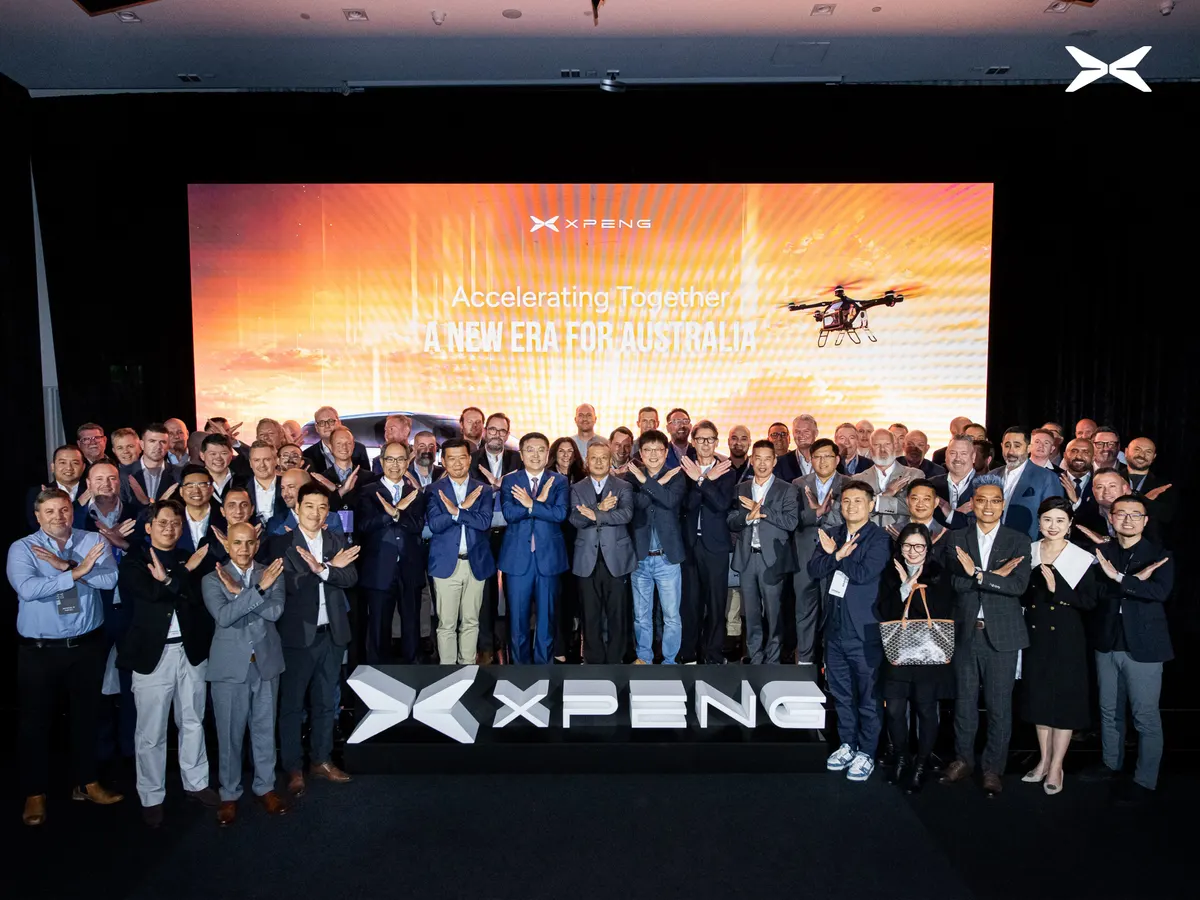 Участники презентации Xpeng в Австралии скрещивают руки в жесте «X» у экрана с дроном и логотипом