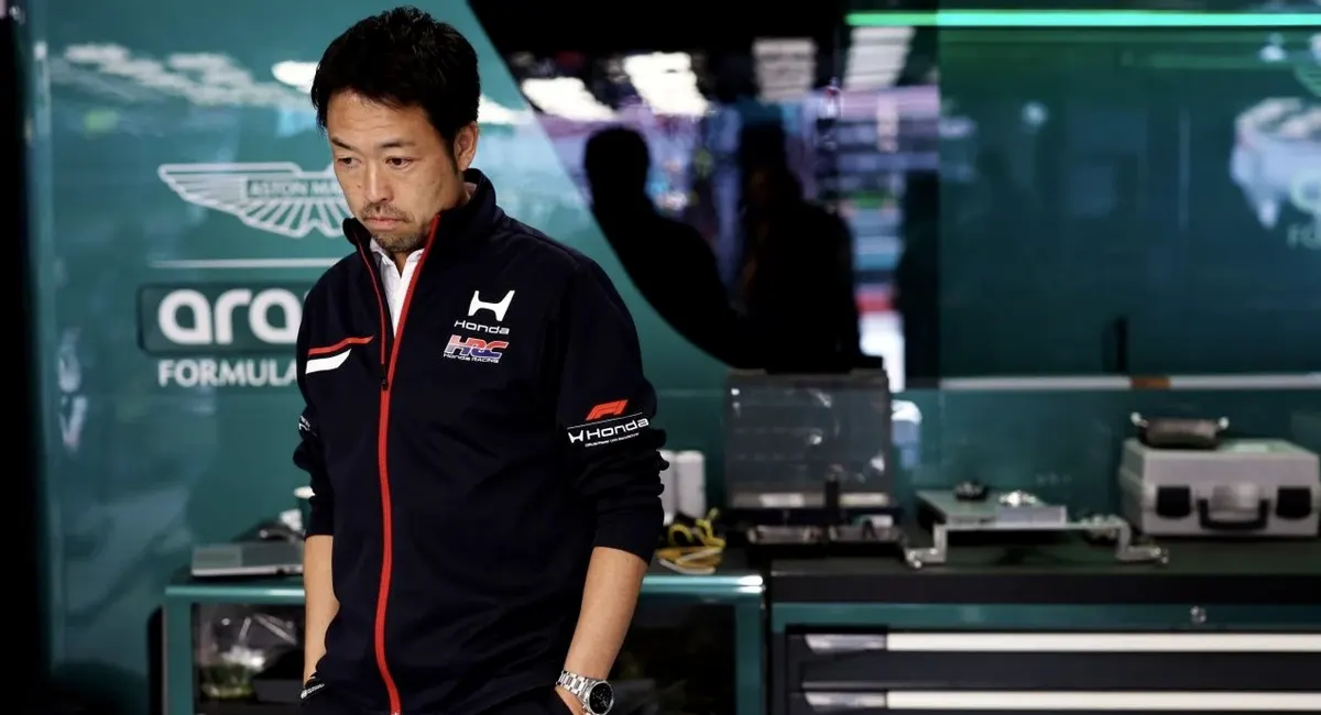 Мужчина в куртке Honda HRC с красной молнией стоит в боксе команды Aston Martin Formula 1, опустив взгляд