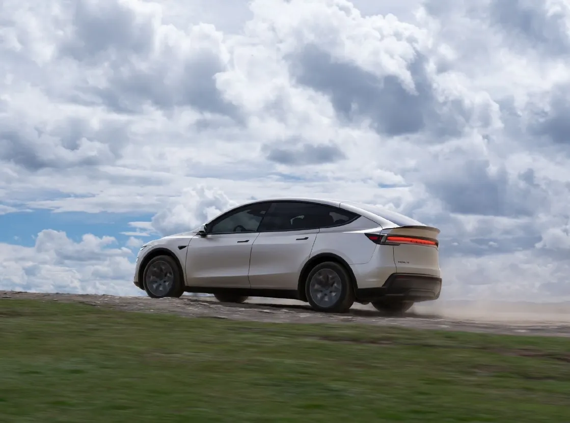 Светлый кроссовер Tesla едет по грунтовой дороге на травянистом склоне под облачным небом, сзади пыль