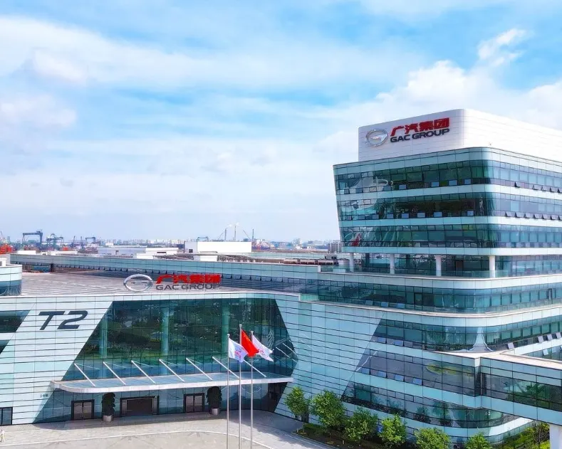 Стеклянное здание с логотипом GAC Group, у входа обозначение T2 и флаги, вдали портовые краны