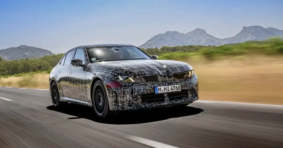 Камуфлированный прототип BMW 3 Series (G50) в движении по трассе на фоне гор, вид спереди сбоку
